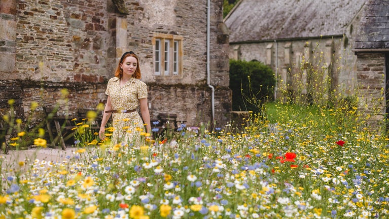 Red, yellow, white and blue wildflowers are slightly out of focus in the foreground. A woman in a short sleeved dress walks behind the flowers, with stone buildings visible in the background.